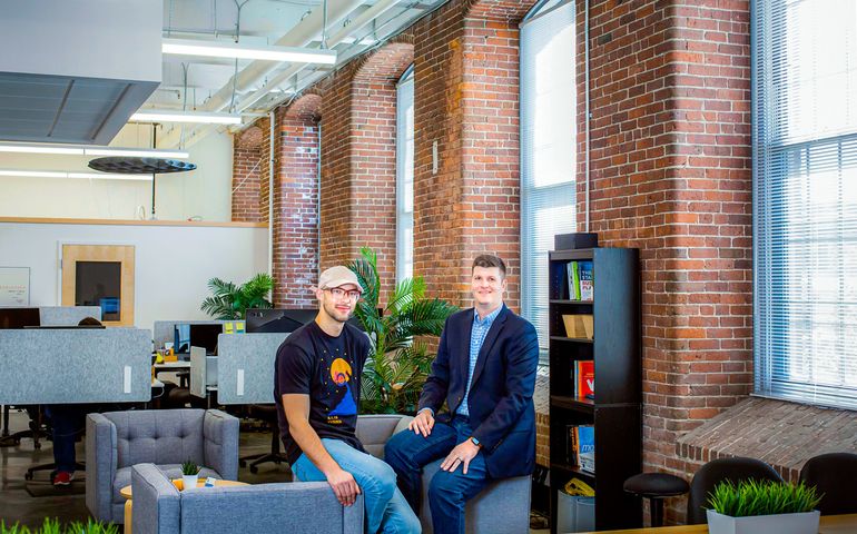 Two men sit on the arms of chairs in an expansive brick-walled working swpace. One is wearing a T-shirt and jeans, the other a a suitjacket, with jeans and a plaid shirt.