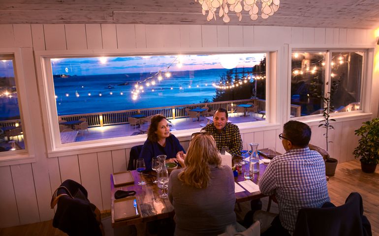six people sit at a restuarant table in front of a large window that shows a harbor at dusk
