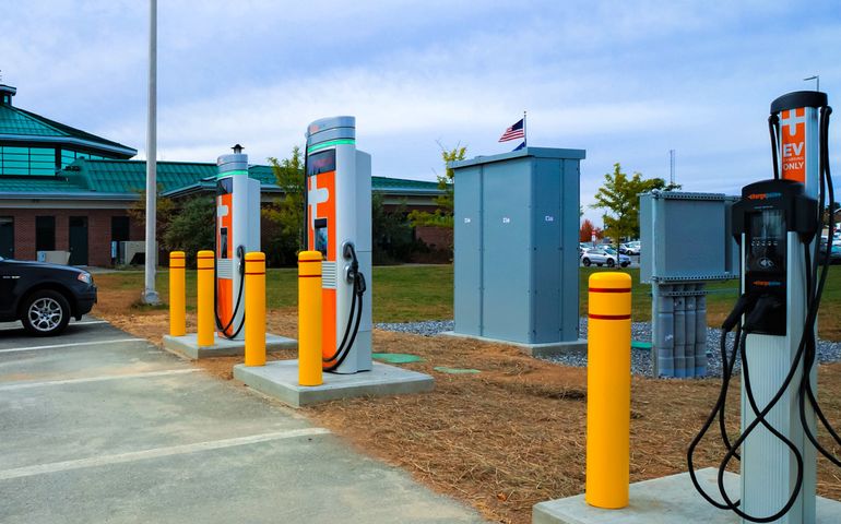 two high speed charging stations that are white with bright orange features and a glimpse of the west gardiner travel plaza in the backgroundtion