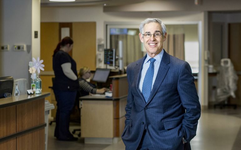 a smiling white man with white hair and glasses in a blue suit stands in a hospital corridor